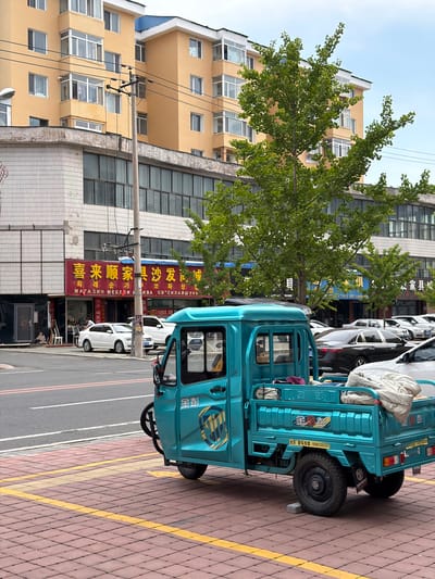 In Hunchun, China, a turquoise three-wheeled vehicle is parked on a red brick sidewalk with yellow tactile paving lines. The vehicle has a covered bed with a light-colored blanket draped over it. The side of the vehicle features a logo with stylized text and a shield emblem. In the background, a multi-story yellow building with balconies and numerous windows is visible. Adjacent to this building is a lower commercial structure with large glass windows and signage in Chinese characters, indicating a furniture and upholstery store. Several cars are parked along the street in front of the commercial buildings. A utility pole with electrical wires stands near the center of the frame, and green trees with lush foliage are present on the right side of the image. The street in front of the sidewalk is paved and appears to be a road with lane markings.