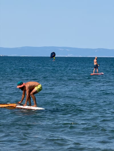 The image captures a sunny day at the sea in Atiya, Bulgaria, with several individuals engaged in water sports. In the foreground, a man wearing a green hat and yellow shorts is crouched on a paddleboard, appearing to adjust equipment. Further out in the water, another man stands on a paddleboard, while a third person is visible in the distance on a paddleboard with a wing sail. The sea is a deep blue, with moderate waves, and a distant coastline with buildings and hills is visible under a clear blue sky.