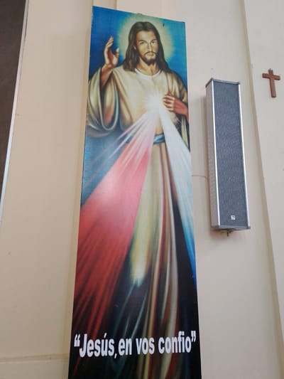 A vertical photograph depicts a religious image of Jesus Christ, displayed on a wall next to a speaker and a wooden cross. The image of Jesus, with long brown hair and a beard, is painted in a traditional style with rays of light emanating from his chest. Below the image, the words "Jesús, en vos confío" are written in Spanish. The speaker is tall and rectangular, mounted on the wall. A small wooden cross is attached to the wall to the right of the speaker. The background is a plain wall, likely inside a church. The location is Villa Hayes, Paraguay.