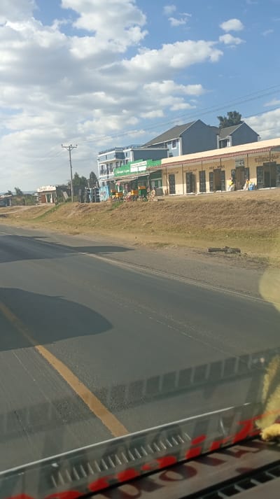 A roadside view shows a two-story building with blue and white accents and a lower, connected structure of tan storefronts, including “LAOZI GEM SHOP”. A row of vendor carts lines the sidewalk. The road stretches toward the horizon under a sky dotted with clouds. A yellow dividing line marks the asphalt. The image appears to be taken from inside a vehicle, with parts of the dashboard visible.