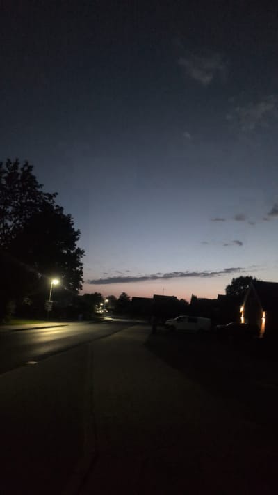 The image depicts a street scene at dusk in Hetlingen, Germany. The sky displays a gradient of dark blue to a faint orange and pink hue near the horizon, with scattered clouds. On the left side of the image, a silhouetted tree and a street lamp illuminate a portion of the asphalt road. The road curves into the distance, lined with dark silhouetted houses on the right. A white van is parked on the right side of the road, with another vehicle partially visible behind it. The overall impression is a quiet residential street as evening sets in.