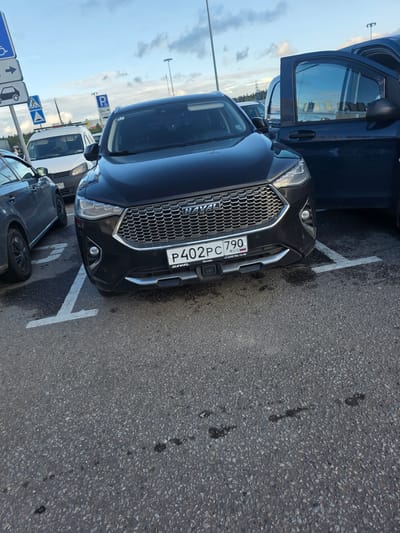 This image shows a black Haval SUV parked on asphalt in Sholokhovo, Russia. The car is angled, with its front bumper and grille prominently displayed. The license plate is visible and reads "P402PC 790 RUS". Text on a sticker below the license plate indicates "+7 (495) 956-13-00" and "Москва".
Other vehicles are visible in the background, including a dark blue SUV with its door open and a white van. Several road signs are also present, including parking signs with a blue "P" symbol and arrows, and a pedestrian crossing sign. The sky is overcast with some clouds, suggesting it might be daytime but not necessarily sunny. The scene appears to be a parking lot, likely in a public area given the presence of multiple vehicles and signage. There are no people visible in the image. The asphalt shows some dark stains, possibly oil or other fluid leaks. The overall impression is a utilitarian, everyday scene.