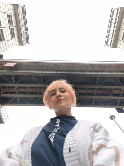 An extreme low-angle, eye-level, medium shot captures a young woman with short, dyed blonde hair standing under a bridge. She is looking directly at the camera with a neutral expression. She is wearing a dark blue t-shirt with white lettering and a cream-colored cardigan with a brown heart pattern. The bridge above her is made of concrete and metal. Two buildings with multiple windows are visible in the upper corners of the frame, suggesting an urban environment. The sky is overcast and white. The scene is situated in Vytegra, Russia.