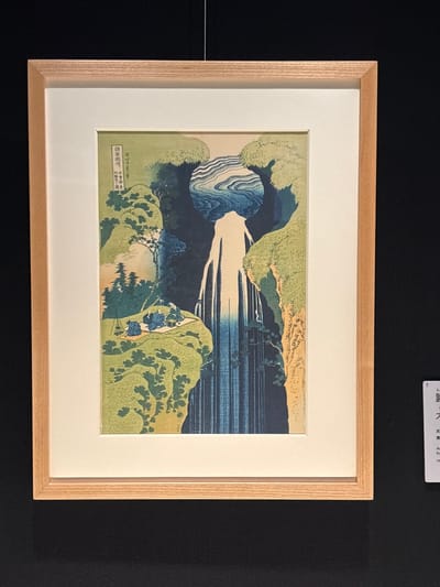 This is a photograph of a framed woodblock print hanging on a black wall. The print depicts a landscape with a waterfall. The waterfall is central to the image, cascading down into a dark, vertical crevice. At the top of the waterfall, a circular pool of water with swirling patterns is visible, framed by rocky outcrops and lush green foliage. In the mid-ground, to the left of the waterfall, several figures are seated on a flat, grassy area. They appear to be gathered around what looks like a picnic, with some objects on the ground that could be food or supplies. The figures are small and indistinct, wearing dark clothing. Trees and dense green vegetation cover the hillsides. The overall color palette is dominated by shades of green, blue, and brown, with some hints of yellow and orange in the landscape. In the upper left corner of the print, there are vertical lines of text in Japanese characters. To the right of the framed print, a small white placard with Japanese and English text is visible. The placard appears to contain information about the artwork, possibly its title, artist, and origin. The print is framed in a light-colored wooden frame with a white mat.