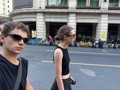 The image depicts an urban street scene featuring two young adults, a woman on the left and a man on the right, both wearing black sunglasses. The woman has light brown hair tied back, is dressed in a black sleeveless crop top and dark shorts, and holds a smartphone. The man has brown hair and wears a black t-shirt with a dark shoulder bag strap across his chest. Both individuals are looking forward or slightly down, suggesting they are walking or paused on a city sidewalk.

The foreground and mid-ground show an asphalt street, which appears wet in areas, indicating recent rain or overcast conditions. A yellow median line and white road markings are visible. Along the edge of the street, a long row of shared bicycles, including yellow and blue/black models, are neatly parked.

In the background stands a multi-story building with a distinct architectural style, featuring prominent vertical pilasters, arched sections, and multiple windows. Some windows are dark, while others have decorative awnings. A small yellow sign with Chinese characters is affixed to one of the building's columns. Reflected in a dark window, reversed text resembling "Bolong" is visible, and a partially obscured red sign on an adjacent building also displays Chinese characters. The scene is set during daytime, with soft lighting.