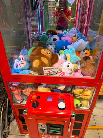 The image shows a brightly lit interior of a claw machine filled with plush toys. A woman wearing a red shirt is visible in the reflection of the glass, holding a camera. The claw machine is predominantly red with black accents and has a transparent front where the toys are displayed. Various stuffed animals, including a large brown bear, a blue monkey-like character, and a white bunny with pink accents, are piled inside.
Attached to the glass of the machine are several stickers with text in Russian. One sticker prominently displays the word "ВНИМАНИЕ!" (Attention!) and provides instructions regarding payment methods, mentioning "monetary denominations" and "coins". Another sticker, with the heading "ПРИЛОЖИТЕ КАРТУ ИЛИ ТЕЛЕФОН" (Tap card or phone), indicates the cost of "4 attempts" for "80 rubles". There is also contact information for technical support with a phone number and Telegram/WhatsApp details.
The scene appears to be indoors, likely in a public place like a shopping mall or arcade, given the nature of the machine and the surrounding environment, which is partially visible in the reflections and the background. The lighting is artificial and focused on the toys, creating a vibrant and playful atmosphere. The overall impression is of a typical amusement or entertainment setting.