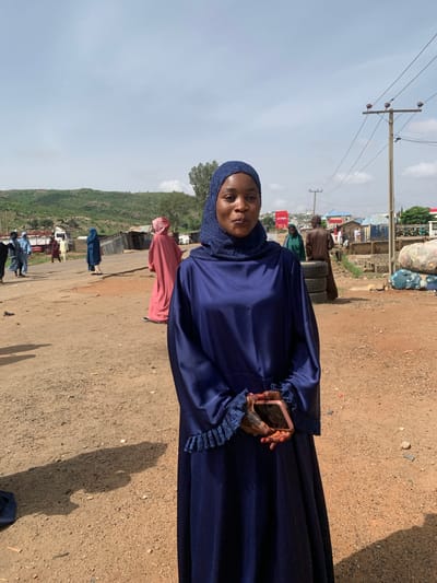 A woman wearing a blue hijab and a long blue dress is standing on a dirt road in Naraguta Hausawa, Nigeria. She is holding a pink cellphone in her left hand. In the background, there are other people walking along the road and some buildings. There is also a green hill on the left side of the road. A row of telephone poles can be seen on the left side of the road. The sky is partially cloudy.
