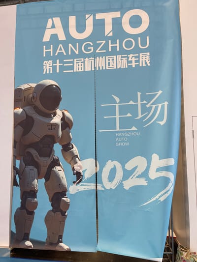 The image shows a large banner for the "Hangzhou Auto Show 2025" located at the Hangzhou International Expo Center in Hangzhou, China (30.272, 120.416). The banner features a large robot in a white and gray suit, standing in front of the words "Auto Hangzhou" and "主场" (meaning "Main Field" in Chinese), and the year "2025" written in white on a light blue background. The image depicts a promotional banner for the upcoming auto show. The banner is displayed outdoors in a public area. It is likely part of a larger promotional campaign to draw attention to the event.