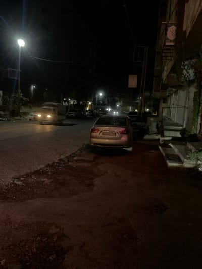 A narrow, dark street at night, lined with buildings, shows a silver car parked on the right. Streetlights illuminate a line of parked cars further down the road. The street surface is rough. The sky is black. The buildings are tall and close together, with greenery growing on some. The scene is poorly lit, adding to the nighttime atmosphere.