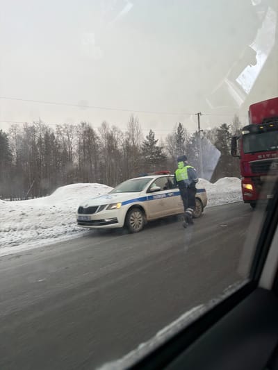 The image depicts a winter roadside scene in Yus'ki, Russia. A white and blue Russian police Skoda sedan, marked with "ПОЛИЦИЯ" (POLITSIA) on its side and displaying active red and blue emergency lights, is stationary on the right side of a snow-dusted asphalt road. The vehicle's license plate, "M276TA 777RUS," is visible. A male police officer stands near the driver's side of the police car, wearing a dark uniform, a dark winter hat, and a highly visible neon yellow-green reflective vest. His left arm is slightly bent, and his right arm is mostly obscured. To the immediate right of the police vehicle, the front right portion of a red Sitrak semi-truck with illuminated headlights is visible, partially obscured by the interior frame of the vehicle from which the photograph was taken. The environment features snow-covered ground, forming banks along the road, and a dense forest of bare deciduous and evergreen trees in the background under an overcast grey sky. Power lines and a utility pole are also visible in the middle ground. The road surface appears wet or slushy, indicating cold conditions.