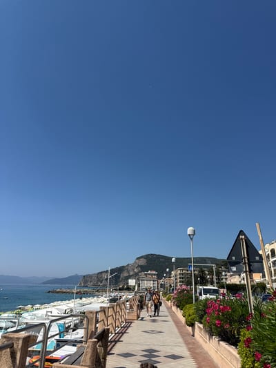 This image depicts a sunny day in Finale Ligure, Italy, along the beachfront promenade. The clear blue sky dominates the upper half of the frame, with a faint crescent moon visible. The promenade is paved with patterned tiles and lined with a low stone wall and planters overflowing with vibrant pink and green foliage. Several streetlights are visible along the promenade.

In the middle ground, three people are walking along the promenade. One man and two women, all dressed in casual summer clothing, are walking towards the left side of the image. A white van is parked to the right of the walkway, behind a flower bed.

Further in the background, the coastline of Finale Ligure is visible, with a series of buildings nestled at the base of a rugged, green-topped mountain. The sea stretches out to the horizon, dotted with a few small boats. On the left side of the image, numerous white and yellow beach umbrellas and lounge chairs are set up on the beach, indicating a popular tourist destination. The overall scene suggests a pleasant summer day at a seaside resort.