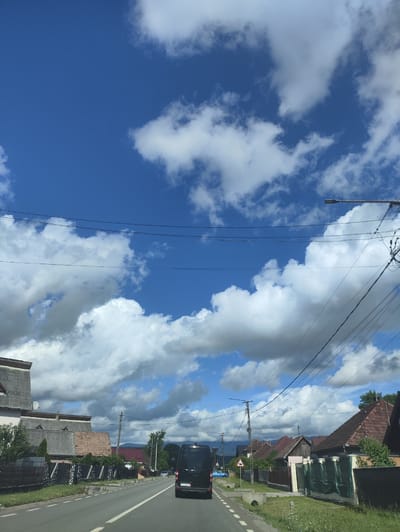The image shows a view from a vehicle driving down a rural road in the city of Crăciunești, Romania. The sky is a vibrant blue with scattered white cumulus clouds. On the left side of the road, there are houses with grey roofs and some greenery. On the right side, there are more houses with red tile roofs and fences. A black van is driving in front of the vehicle. The road has white lines marking the lanes. Telephone poles and wires are visible along the side of the road. In the distance, mountains can be seen. The overall scene depicts a typical day in a Romanian village.