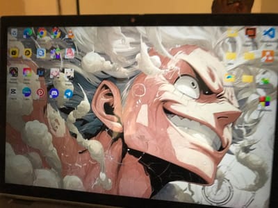 The image displays a laptop screen in an indoor setting within the city of Calabar, Nigeria. The screen features a desktop background dominated by an anime-style illustration of a character's face, characterized by spiky white hair, a wide, exaggerated grin with sharp teeth, and a manic expression. The character's skin is a reddish-pink hue, set against a backdrop of white and grey cloud-like formations. The desktop is populated with various application icons, including Google Chrome, VLC media player, Adobe Photoshop, Adobe Lightroom, Discord, Telegram, Pinterest, Canva, and Genshin Impact. Minor scratches are visible on the screen's surface. Above the screen, a blurred reflection, likely of a dark-skinned individual's head with eyeglasses, is partially discernible against an out-of-focus, light-colored background.