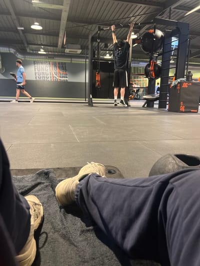 The scene depicts the interior of a fitness center, located in Annœullin, France. The perspective is from a low vantage point, focusing on the foreground with an individual's legs and feet resting on a dark grey towel on the floor. These legs are clad in navy blue sweatpants and light-colored athletic shoes. A black, possibly cast-iron, gym weight is partially visible to the right of the foreground subject.

In the midground, a male individual is observed from the rear, suspended from a black pull-up bar structure, with his arms extended upwards, grasping the bar. He wears a dark short-sleeved shirt, black shorts with white vertical stripes on the sides, and dark athletic shoes with white soles. To his right, within the same black metal rig, are several black spherical weights (likely medicine balls) hanging from orange straps, and a set of orange suspension training straps. Further to the right, a black plyometric box with white numerical markings such as '-20', '-24', and '-30', and the brand 'VARX' is visible.

In the background, a second male individual walks from left to right, wearing a light blue short-sleeved shirt, dark shorts, and light-colored sneakers, holding a dark object in his left hand. Behind him, on a dark grey wall, a graphic displays the word "FUNCTIONAL" repeated in white and orange text. The floor throughout the facility is covered with dark grey interlocking rubber matting. The ceiling features exposed industrial-style ductwork, structural beams, and overhead fluorescent lighting fixtures.