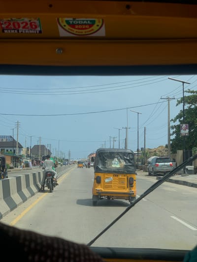 The image shows a view from inside a yellow vehicle, likely a "Keke Napep" (a type of auto-rickshaw), traveling along a road in Satellite Town, Nigeria. The "Keke" has stickers on the left interior, including one that says "2026 KEKE PERMIT" and another that says "TODAL 2026 ORE ADE ZONE". Another "Keke" is visible ahead, with writing in Yoruba "OLA MONEY, OMO IYA" and registration "MUS500PJ" clearly visible. On the right, there's a road divider with a motorcycle parked beside it, and a man in a white and grey striped shirt is working on the bike. Further down the road, there is traffic in both directions and streetlights along the road. There are also power poles with wires. The sky is clear. Buildings and solar panels are seen at the bottom of the image along the side of the road, likely a street market.