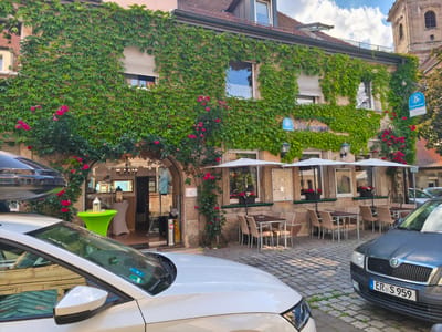 The image depicts the exterior of a building in Erlangen, Germany, likely a restaurant, heavily adorned with lush green ivy and interspersed with vibrant pink roses. The building features a traditional facade with windows and shutters, and a tiled roof. In the foreground, a white car is parked, and to its right, a dark gray car with a license plate reading "ER S 959" is visible.
The ground is paved with cobblestones, and there are outdoor seating areas with tables and chairs set up under umbrellas, suggesting a cafe or restaurant. Through an arched entrance, the interior of the establishment is partially visible, with tables, chairs, and decorative items. A bright green round table with a lantern on top is positioned near the entrance.
A prominent blue sign with white lettering, likely indicating the name of the establishment, is attached to the building. The overall scene suggests a pleasant outdoor dining or seating area in a European city. The presence of the "ER" prefix on the license plate confirms the location as Erlangen.