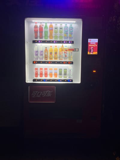 A brightly illuminated beverage vending machine stands prominently in a dark, outdoor urban environment in Yanqiao, China, likely at night. The machine's transparent front displays three rows of various bottled and canned drinks, including Coca-Cola, Fanta, Sprite-like beverages, fruit juices, and Monster Energy drinks. Several display sections show "5°C," indicating the temperature at which the drinks are held. Below the main display, a red rectangular sign features the Coca-Cola logo in white Chinese characters ("可口可乐"). To the right of the vending machine's primary display, a smaller vertical digital screen advertises "COSTA COFFEE" with a price of "2元," and a small amber indicator light is visible below it. The surrounding area is largely dark, with faint, blurry blue and purple light sources and indistinct features visible in the background, including a partial number sequence that appears to be "22045494." No individuals, active interactions, or discernible motion are present in the scene.