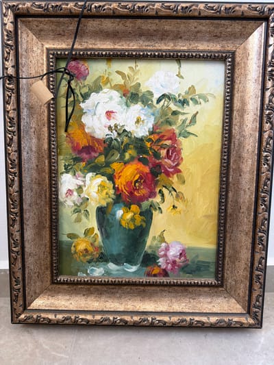 The image shows a framed painting of a floral still life, likely a bouquet of roses in a vase, with a vibrant yellow background. The painting is in portrait orientation and is enclosed in an ornate, textured wooden frame with a golden-brown hue. A black wire with a small, light-colored cylindrical object attached is draped over the top left corner of the frame, partially obscuring the painting. The painting itself appears to be executed in an impressionistic style with visible brushstrokes. The floral arrangement features a variety of rose colors, including white, red, orange, and yellow, with green foliage. The vase is a dark teal or green color. The overall impression is one of a traditional still life artwork. The location is specified as Sderot, Israel, but the image itself does not provide any direct visual cues to confirm this specific location within the city, such as landmarks or specific urban features. The setting appears to be indoors, with the framed artwork placed on a flat surface that resembles a tiled floor.