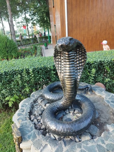 The image shows a snake sculpture in the foreground, with a lush green hedge behind it. A wooden building is visible to the right. In the background, a park setting is depicted with trees, flowers, and several people. A child in a white hat is visible near the wooden building. The location is identified as Kenbulun, Kyrgyzstan.