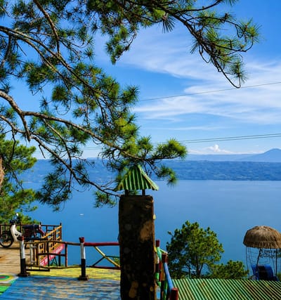 This is a landscape image taken from a high vantage point overlooking a large body of water, likely Lake Toba, given the context of Parik Sabungan, Indonesia. The foreground features a wooden deck with colorful painted planks and a railing system composed of wooden posts painted in red, white, and yellow. On the deck, a red and white motorcycle is parked. To the right of the deck, a stone pillar stands topped with a miniature green, corrugated metal roof, resembling a small chimney. Further to the right, a thatched-roof seating area is visible. Dense green pine trees are scattered throughout the scene. The middle ground is dominated by the vast, calm, blue expanse of the lake, with distant hills and mountains forming the horizon. The sky is a clear blue with wispy white clouds. The image is captured on a sunny day, with the sunlight illuminating the scene and casting shadows.
