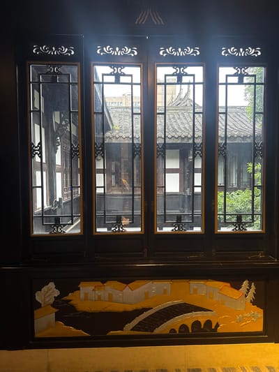 This image captures an interior view looking out through a traditional dark wooden window in Qingyang District, China. The window structure consists of four vertical panels, each featuring a grid of dark, ornate bars and lighter brown/orange frames around clear glass panes. Above each pane section, the dark frame is adorned with white, intricately carved scrollwork. The view through the window reveals traditional Chinese architectural elements: several grey, tiled rooftops with undulating patterns and decorative ridges. Beyond these roofs, glimpses of white and dark wooden building walls, balconies, and other window frames are visible, suggesting a courtyard or narrow street environment. Green foliage from trees or shrubs is apparent through the rightmost panes. In the upper background, faint outlines of taller, more modern buildings can be seen above the traditional structures. The interior space, where the photo is taken from, is dimly lit, silhouetting the window frame against the brighter exterior. Below the windowpanes, a dark horizontal panel features a three-dimensional relief or painting depicting a landscape scene. This scene includes several white houses with golden roofs, golden landmasses, a dark river or path, and a multi-arched silver bridge. A white, stylized tree is on the left, and two evergreen trees are on the right side of this decorative panel. No individuals are clearly discernible in the scene.