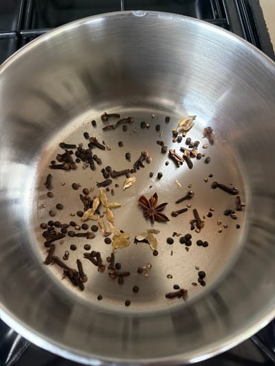 A top-down, close-up shot captures a collection of whole spices, including star anise, cloves, cardamom pods, and peppercorns, scattered loosely within a brushed stainless steel pot. The pot is placed on a black stovetop, with the edge of the stovetop visible in the upper portion of the frame. The lighting appears to be overhead, casting gentle reflections on the metallic surface of the pot. The image offers a clear view of the textured surfaces and distinct shapes of the individual spices.