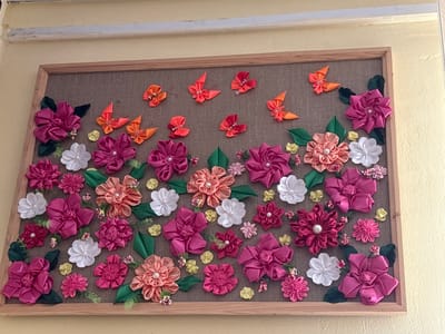A framed textile art piece is displayed on a wall. The art piece features numerous fabric flowers and butterflies in shades of pink, orange, red, and white, arranged on a brown fabric background. The frame is made of light-colored wood. The wall appears to be painted a pale yellow. The image is taken from a slightly elevated, frontal perspective. The scene does not show any people.