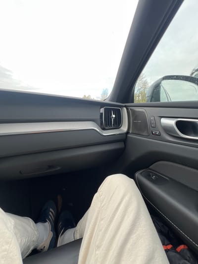 The image displays the interior of a vehicle from the perspective of a passenger, looking forward and slightly right, located in Villarboit, Italy. The dashboard is dark grey, featuring a horizontal silver trim element and a chrome-accented air vent on the right side. The passenger door panel, also dark grey, includes a "Harman Kardon" branded speaker grille, three memory seat control buttons, and a silver door handle. The lower section of a person's legs, clad in light beige or off-white pants, are visible in the foreground. The person's feet, wearing dark blue athletic shoes with white stripes and white socks, rest on the black floor mat of the passenger footwell, which contains small pieces of debris. Through the windshield and passenger window, an overcast sky is visible above a distant treeline. A dark vehicle is partially reflected in the passenger side mirror.