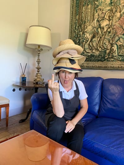 A woman with dark hair is lying on a bright blue leather couch, wearing a white shirt and a dark vest. She has several straw hats stacked on her head, and she is sticking her tongue out playfully at the camera. Her right hand is raised, and she is giving a middle finger. Behind her, a tapestry hangs on the wall, depicting a scene of people on horses. To her left, a white lampshade with decorative trim sits atop a metallic lamp base. A wooden table with a reflective surface is to her right. The setting appears to be indoors, possibly a living room, with tiled flooring visible. The overall mood is humorous and irreverent.