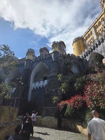 The image depicts the Pena Palace in Sintra, specifically within the city's parishes of Santa Maria e São Miguel, São Martinho e São Pedro de Penaferrim, Portugal. The scene captures a low-angle view of the palace's distinctive architecture under a partly cloudy blue sky.
The central subject is the multi-colored palace, characterized by grey/blue stone walls adorned with crenellated battlements and multiple yellow-capped conical towers. A prominent large yellow cylindrical tower with arched windows is visible on the upper right, connected to a section of the palace with intricate yellow ornamentation. Below this, large dark grey arched structures are visible, one revealing a white crenellated internal structure. The palace walls are partially overgrown with lush green and red-leafed vegetation, including dark green shrubs and bright red bushes along the right side of the paved path.
A group of approximately seven to eight individuals occupies the lower half of the image. They are walking uphill on a light-colored cobbled stone pathway. In the mid-ground, a man in a white short-sleeved shirt and dark shorts stands, facing away from the camera, and gestures with his right arm towards the palace. To his left, a woman in a dark jacket walks beside him. In the immediate foreground, a person in a light grey t-shirt walks away from the camera, partially obscuring other individuals. A dark-clad figure is partially visible on the far right, near the red bushes. The sun illuminates the scene, casting shadows on the pathway and highlighting the palace's vibrant colors against the sky.