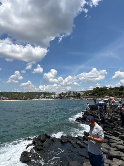 The image depicts a scene at Gành Đá Đĩa, located in the city of Phú Lương, Vietnam. The foreground is dominated by a man wearing a blue and orange baseball cap and a grey polo shirt, looking down at his mobile phone. He is standing on a formation of dark, columnar basalt rocks that extend into the choppy, blue-green ocean. Numerous other people are scattered across the rocks, some holding umbrellas, suggesting a tourist or recreational area. In the background, a coastline stretches out, featuring a sandy beach lined with palm trees and various buildings, including hotels and restaurants. The sky above is a vibrant blue, dotted with fluffy white clouds. The waves are crashing against the basalt formations, creating white foam.