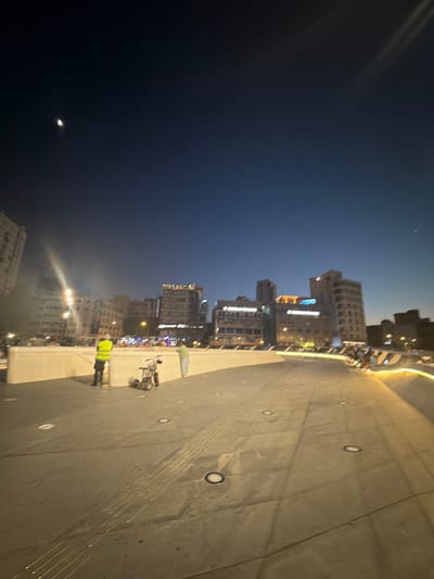 This image depicts an urban scene at night in Nicosia, Cyprus. The orientation of the image is sideways. The foreground is dominated by a textured concrete surface with embedded circular lights and linear markings. 

In the middle ground, two individuals are visible on the concrete path. One person, wearing a bright yellow vest, appears to be standing near a bicycle. Another person, wearing lighter clothing, is seen further away. Beyond them, a series of multi-story buildings are illuminated by numerous lights, suggesting a commercial or residential area. Some of these buildings have prominent signage, though the text is not clearly legible. The architecture suggests a modern urban environment.

The background is a dark night sky. A crescent moon is visible, along with a faint star or distant light. The overall ambiance is one of a city at night, with artificial lights from buildings contrasting with the natural darkness of the sky. The lighting creates a somewhat dramatic and atmospheric effect.