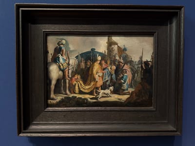 The image shows a framed painting displayed on a blue wall. The painting depicts a biblical scene, likely an Old Testament narrative, with numerous figures engaged in various activities. In the foreground, a white horse with a rider dressed in oriental attire stands on the left. To the right of the horse, a group of people are gathered. One central figure, adorned in a richly patterned gold robe, appears to be interacting with others. A small spotted dog is also present in the foreground, looking towards the right. The background features a large gathering of figures, some carrying weapons, and hints of a tent-like structure and possibly a dark, domed edifice. The painting is set within a dark, wooden frame with multiple layers and a textured surface.
This painting, titled "The Queen of Sheba before Solomon" by Rembrandt van Rijn, is located in the Kunstmuseum Basel, Switzerland. The museum is situated in the city of Basel, near the Rhine River.