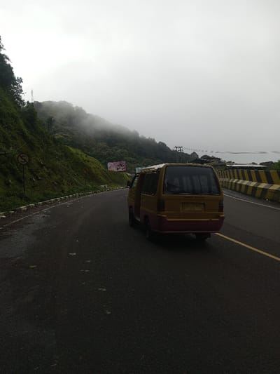 This image captures a two-tone van, primarily yellow/beige with a pink/red lower rear section, on a two-lane asphalt road. The van features darkened rear windows, and the text "Cipanas Puncak" is visible on its upper rear window. The road appears damp and is marked with a single yellow center line. White curb stones delineate the left edge of the road, bordering a steep, densely vegetated hillside covered with trees and foliage, which is partially obscured by thick fog or low clouds. Two circular road signs indicating a curve are positioned on this hillside, and a billboard with an advertisement is visible further up the embankment, partly veiled by mist. On the right side of the road, yellow and black striped concrete barriers separate the roadway from a drop-off. Beyond these barriers, utility poles with wires and a distant body of water are visible through the haze. The sky is overcast, contributing to the foggy and low-light conditions prevalent across the scene. No individuals are visible. While the user specified the location as Bangtsho, Bhutan, the text "Cipanas Puncak" on the vehicle suggests a location in West Java, Indonesia.
