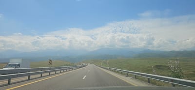 The image captures a view from a moving vehicle on a highway, heading towards a vast mountain range under a bright blue sky with scattered white clouds. The highway has two lanes in each direction, separated by a yellow line. A metal guardrail runs along the right side of the road. To the left, a white box truck is parked on the shoulder, and a yellow and black striped sign is visible. The surrounding landscape is characterized by rolling green hills, some with sparse vegetation and others covered in lush grass. In the distance, the mountains are layered, with softer blues and grays indicating their depth. The scene appears to be a rural or mountainous area outside of a city, possibly near Қырғауылды, Kazakhstan, given the context. The lighting suggests it is daytime, likely mid-morning or afternoon, with the sun casting shadows on the road and hills. There are no visible people in the frame.