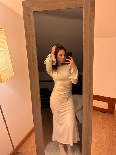The image displays an indoor scene, likely a residential bedroom or dressing area, featuring a female individual captured in a full-length mirror selfie. The specific location within Cava de' Tirreni, Italy, cannot be determined from the visual evidence presented in the image.
The subject, an adult female with dark hair, stands facing the mirror, holding a dark-colored smartphone with a visible multi-lens camera array in her right hand to record the image. Her left hand is positioned in her hair near her head. She is wearing a light-colored, long-sleeved midi-dress that is form-fitting through the bodice and hips, with a boat neck, ruched detailing around the waist, and flared cuffs. Light-colored socks or foot coverings are visible on her feet.
The mirror itself features a wide, light-toned wooden frame with visible grain. To the left of the mirror, a floor lamp with a tall, cylindrical beige fabric shade is present. A white, shaggy rug occupies the floor directly beneath the individual, while the visible flooring outside the rug and in the background reflection is light-colored hardwood. The walls in the reflection are a light, solid color. In the reflection to the right of the individual's lower body, a brown rectangular frame or object is leaning against a wall. The overall lighting is warm, suggesting artificial light sources.