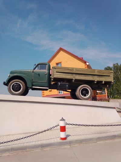 A side view of a vintage green flatbed truck is prominently displayed on top of a low, white concrete wall. The truck is oriented from left to right, with its front wheel on the left. The truck bed is wooden and appears weathered. In the background, an orange brick building with a red roof is visible, featuring windows. To the right of the building, a cluster of green trees is present. A chain barrier with red and white striped posts runs along the front of the concrete wall. The scene is set against a bright blue sky with scattered white clouds. The image appears to be taken outdoors, possibly in a public space or memorial in Krasny Yar, Russia, given the historical nature of the truck and the architectural style of the building.