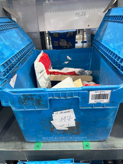 In Nerubais'ke, Ukraine, a blue plastic bin is filled with various parcels and packages. The bin is open and placed on a gray shelf. Several packages are visible, including a large red and white postal bag, a cardboard box, and smaller white envelopes. A white label with a barcode and the text "IBOX-1331272" is affixed to the front of the bin. Another handwritten note with the number "117" is also attached to the bin. The background appears to be an industrial or warehouse setting with metal structures and other blue bins. The overall scene depicts a logistics or sorting area for mail and packages.