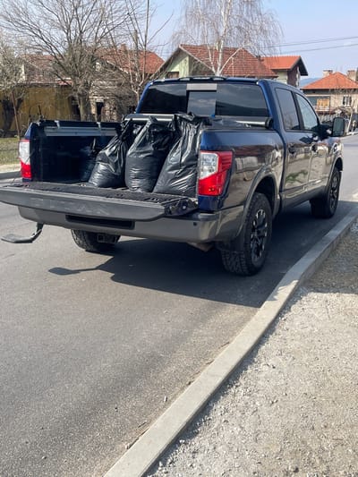 A dark blue Nissan Titan pickup truck is parked on the right side of an asphalt road in Resilovo, Bulgaria, facing away from the viewer. The vehicle's tailgate is lowered, revealing its bed, which contains at least four large, black, opaque bags, appearing full. A deployable step is extended from beneath the tailgate on the driver's side (left from viewer's perspective). The truck features dark-colored rims and all-terrain tires. The red taillight lenses are visible. The road surface is asphalt, bordered by a concrete curb separating it from a gravel shoulder. The scene is illuminated by strong, direct sunlight, casting distinct shadows. In the background, several residential houses with red-tiled roofs are visible amidst bare deciduous trees, indicating a late autumn, winter, or early spring season. No individuals are clearly discernible in the immediate vicinity of the truck.