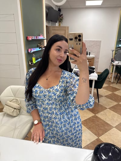 A woman is taking a selfie in what appears to be a nail salon in Kąty Wrocławskie, Poland. She is wearing a blue and white floral dress and has long dark hair. She is holding a gold-colored smartphone in her right hand, which is decorated with various charms. Her left hand is resting on a white surface, revealing a watch on her wrist and a red string bracelet. Next to her, on a white chair, is a small quilted handbag. In the background, shelves are filled with nail polish bottles, and there are salon chairs and tables, along with a tiled floor.