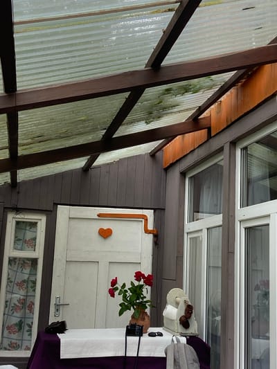 The image shows a covered outdoor area, likely a porch or sunroom, with a corrugated plastic roof supported by wooden beams. The walls are made of dark vertical wooden planks. To the left, a white-framed window with a floral curtain is visible. In the center, a white door with a silver handle is decorated with an orange heart-shaped ornament and an orange hook-like fixture above it. A table covered with a purple tablecloth and a white runner is set up in front of the door. On the table, there are red roses in a clay pot, a black camera, sunglasses, and a vintage-looking grinding mechanism with small woven baskets attached. To the right, a large white-framed window reveals a reflection of the interior, including what appears to be a person or mannequins dressed in white. A gray jacket or bag is draped over the side of the table. The scene appears to be located in the Ратайчыцкі сельскі Савет, Belarus.