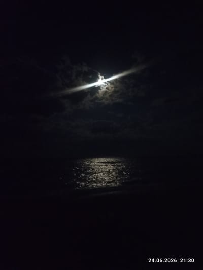 The image captures a nighttime scene at a body of water, likely the Caspian Sea given the location context of Shubatobe, Kazakhstan. The moon is prominently featured in the sky, partially obscured by clouds, casting a bright, shimmering reflection on the dark water's surface. The light from the moon creates a path of glinting highlights that extends across the water towards the viewer. The overall mood is serene and tranquil, with the darkness of the night contrasted by the bright moonlight. The time stamp in the lower right corner indicates the image was taken on June 24, 2026, at 21:30, suggesting it is evening or early night. There are no visible people or man-made objects, focusing the scene on the natural elements of the moon, sky, and water. The weather appears clear enough for the moon to be visible, though clouds are present. The location is implied to be near a coast or large body of water.