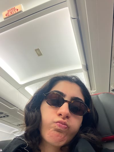 A woman with dark wavy hair and tortoiseshell sunglasses makes a duck face while sitting on an airplane. An illuminated "SAÍDA EXIT" sign is visible on the ceiling of the plane's cabin. The image appears to be taken inside an airplane cabin, possibly during a flight. There is no indication of the location being in Moreira, Portugal.