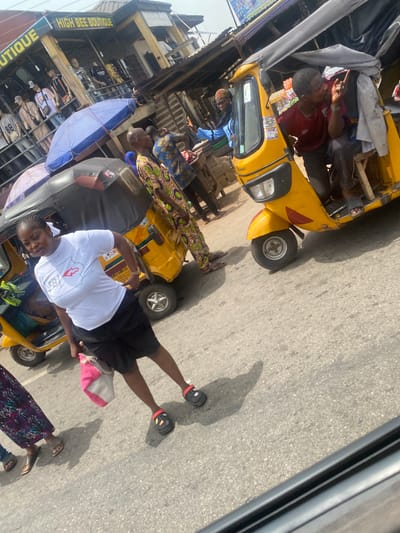 The scene shows a street in Alakija, Nigeria, with yellow auto rickshaws (tuk-tuks) being used for transportation. Several people are present; some are passengers riding in the tuk-tuks, while others are standing nearby on the street. A multi-story building with the sign "HIGH BEE BOUTIQUE" is visible in the background. The setting is outdoors on a sunny day.