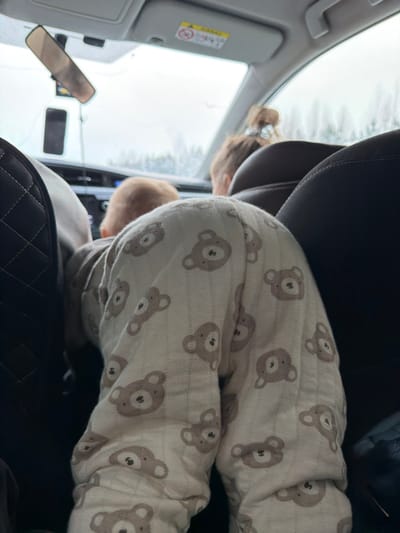 The scene is captured inside a vehicle in the city of Yus'ki, Russia, viewed from a rear passenger seat looking forward. In the immediate foreground, an infant or young child is positioned on hands and knees, facing the front of the car. The child is wearing a light-colored full-body garment patterned with multiple brown bear faces. To the left, a portion of a dark, quilted seatback is visible. To the right, a plain dark seatback is present. Further forward, the heads of two individuals are visible from behind; one with light brown hair in the front passenger seat, and another in the driver's seat with light brown hair tied back. Through the front windshield, an overcast sky and blurred green-brown foliage are visible. A rearview mirror hangs from the windshield, and a dark smartphone is mounted below it. A sun visor with an "AIRBAG" warning label is also visible above the windshield.