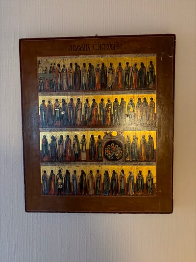 The image shows a religious icon hanging on a textured, off-white wall. The icon is rectangular with a dark brown wooden frame. The icon itself is predominantly gold, with multiple rows of painted figures. The top row depicts numerous figures standing in what appears to be a procession or a gathering. Below this, there are three more rows of figures, each row containing a dozen or more individuals depicted in religious attire. The central area of the third row features a scene with figures gathered around a circular element, possibly representing a biblical event or a saintly depiction. The artwork is detailed, with individual figures painted with distinct features and robes. Some of the figures appear to be holding scrolls or books, and they all have halos around their heads, indicating sainthood or religious significance. The overall impression is that of an Orthodox Christian icon. The text at the top of the icon is in Cyrillic script. This object is located in Osnabrück, Germany.