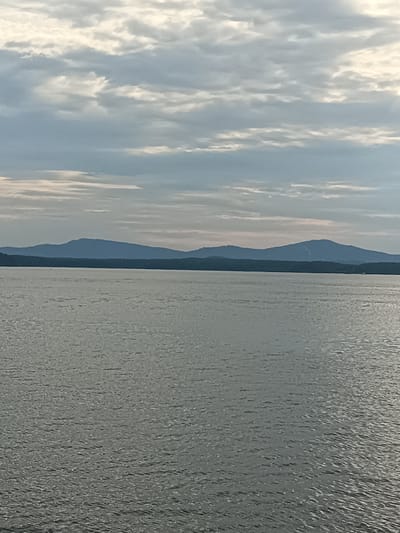 The image depicts a wide expanse of calm water in the foreground, reflecting the overcast sky. In the mid-ground, a dark, tree-lined horizon separates the water from the background, where a range of distant, hazy blue mountains stretches across the frame. The sky is filled with a mix of grey and white clouds, with some patches of lighter sky visible. The scene appears to be a natural landscape, likely a lake or a large river with a mountainous backdrop. The location is described as being in the city of Tatysh, Russia, suggesting a geographical setting with these features.