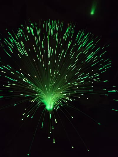The image displays a cluster of green illuminated fibers against a black background. The light source appears to originate from the center, with the fibers radiating outwards in various directions. The fibers have varied lengths and different intensities of green light along their lengths, with brighter points at the ends. There are no discernible people, subjects, objects, actions, or interactions besides the illumination. The environment is completely black except for the light emitted by the fibers. There is no indication of the image being taken in Cherkovitsa, Bulgaria.