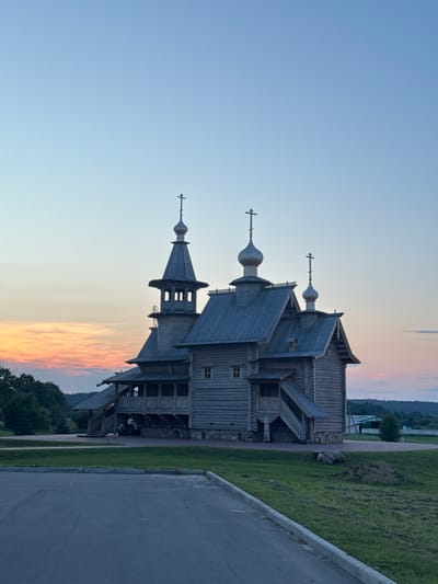 The image shows a wooden church with multiple domes topped with crosses, set against a twilight sky with a colorful sunset. The church is constructed from logs and features intricate wooden carvings and detailing. It is surrounded by a green lawn, with a paved area in the foreground. Trees and a rolling landscape are visible in the background. The location is identified as a church in the Neman rural council, Belarus. There are no people visible in the image.