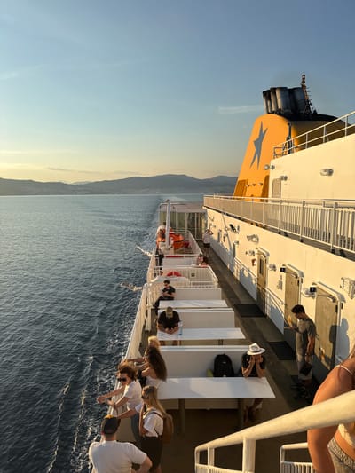 The image depicts a ferry moving through the Aegean Sea, with the coast of Kavala, Greece, visible in the background. The time of day appears to be late afternoon or early evening, with the golden light of the setting sun illuminating the scene.
Several people are visible on the deck of the ferry, some sitting on benches and others standing and looking out at the sea. The mood seems relaxed and peaceful, with individuals enjoying the journey and the scenic views. One person is wearing a white hat and appears to be on their phone. Another group of people is gathered near the railing, observing the water.
The ferry itself is white with yellow accents and features a prominent blue star logo. The sea is a deep blue, with gentle waves indicating a calm day. The coastline is mountainous and appears to be sparsely populated. No visible text is present in the image.